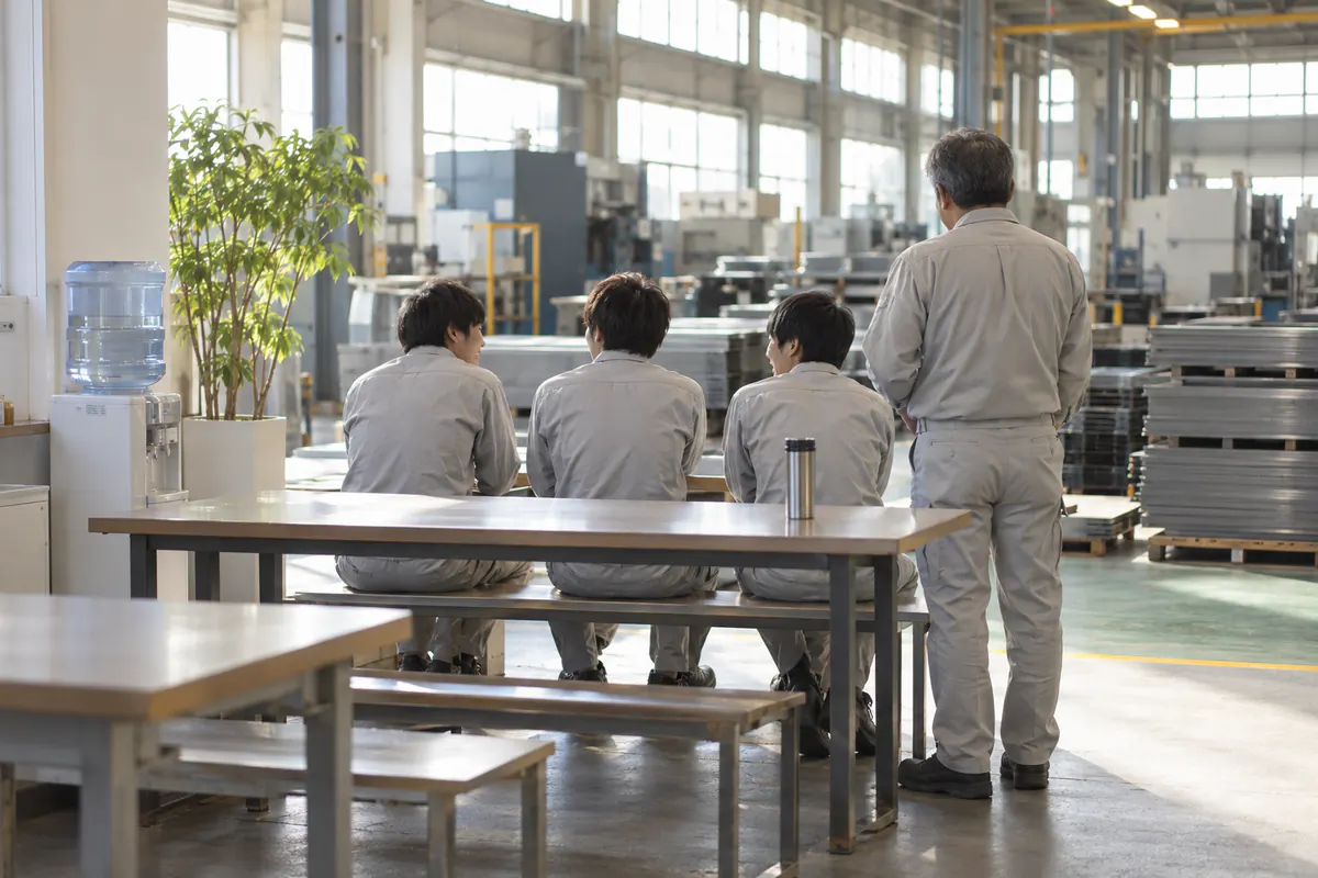 明るい板金工場の休憩スペースで、作業服姿の若手3人がベンチに並んで座って話し、そばに先輩が立っている後ろ姿